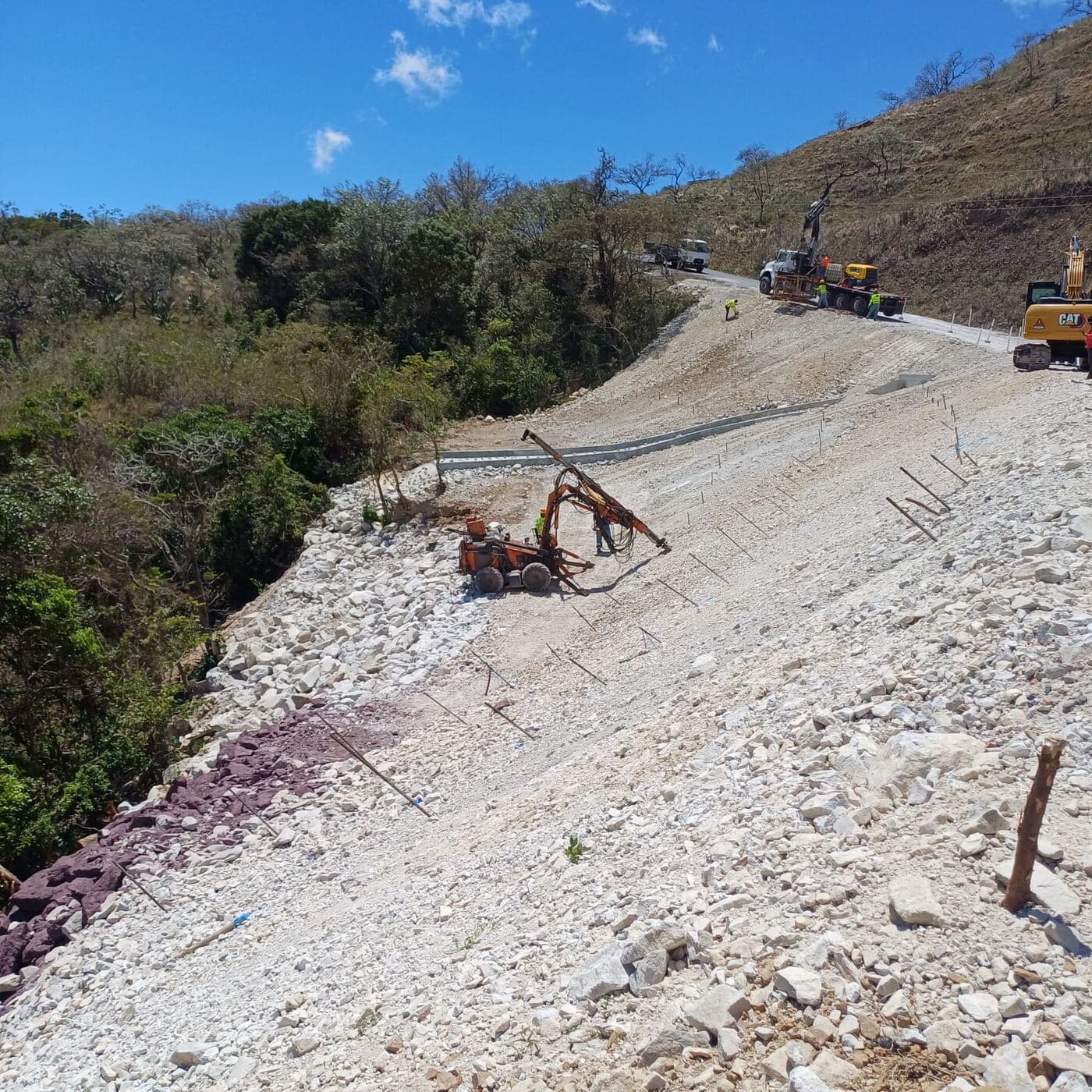 Equipo de perforación instalando anclajes sobre el talud, con filas de anclajes ya colocados y maquinaria en la vía